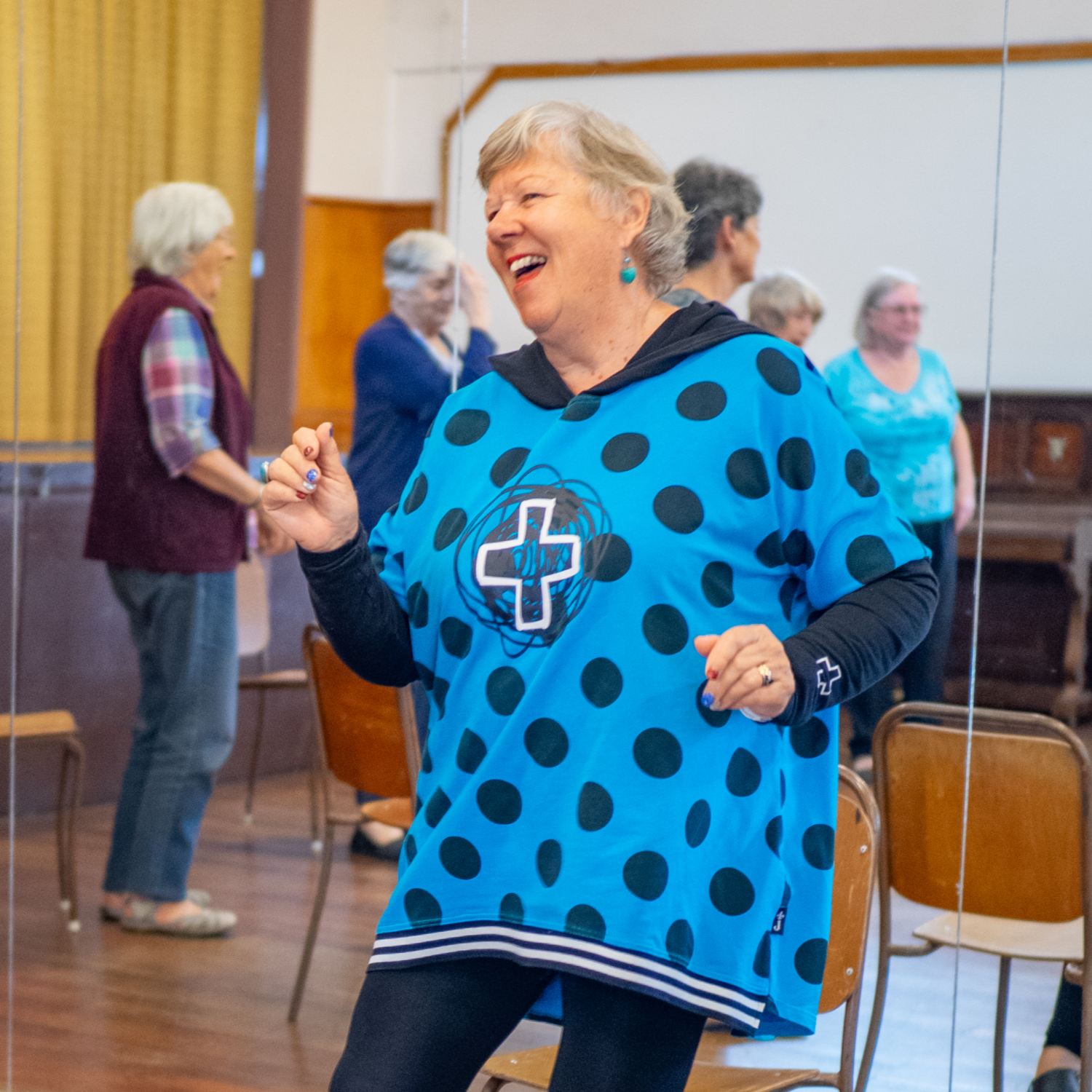 a smiling Pakeha woman in a blue top with black spots is dancing in three quarter profile